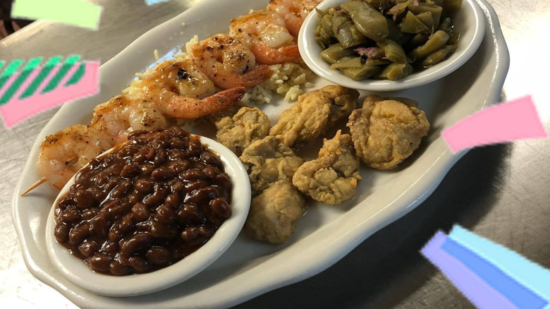a plate of baked beans, fried oysters, grilled shrimp over rice, and green beans on a silver table