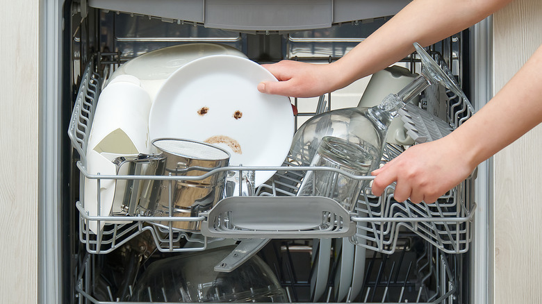 A hand putting dirty dishes into a dishwasher, with a sad face wiped onto a white place