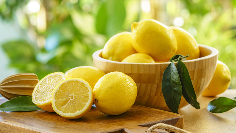 Lemons in a bamboo bowl with a wooden cutting board and citrus reamer on a table outside.