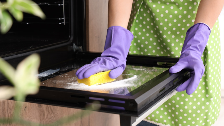 A person in a green polka dot apron with purple gloves cleaning an oven door with a yellow sponge
