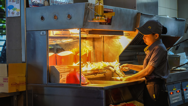 A McDonald's employee wearing a hat filling a french fry container in a kitchen.