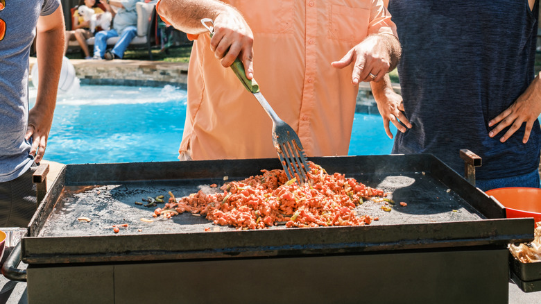 Three men cooking taco meat on a griddle with a pool in the background.