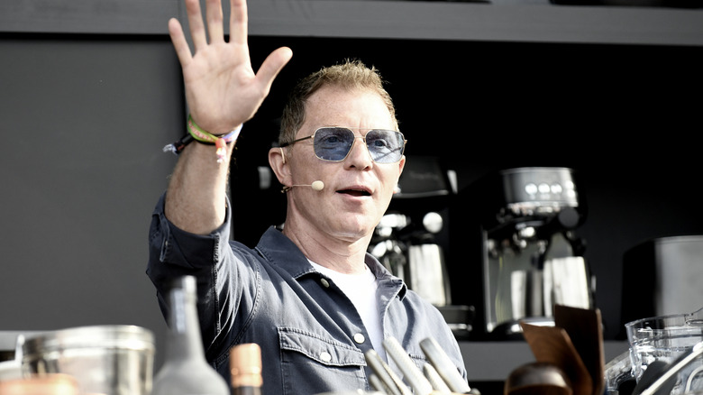 Bobby Flay raising his hand while talking on a stage with blue sunglasses on