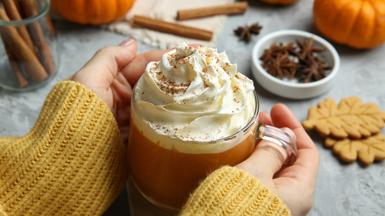 Hands holding a glass mug of pumpkin spice latte topped with whipped cream and spices with blurred anise, cinnamon sticks, and orange pumpkins in the background