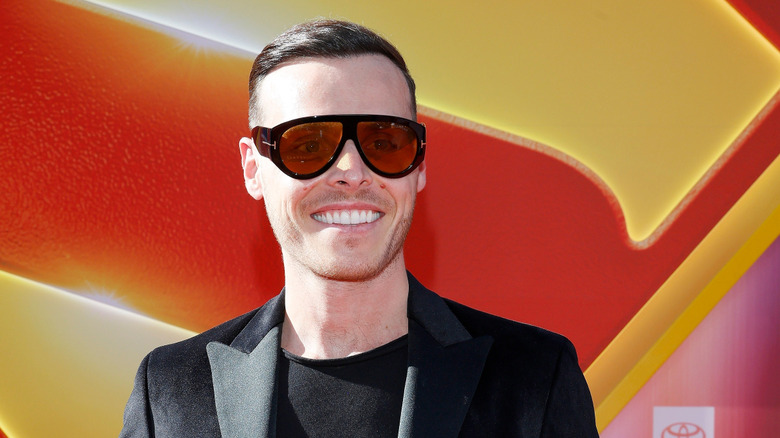 Amaury Guichon in large brown sunglasses, black suit, and black t shirt