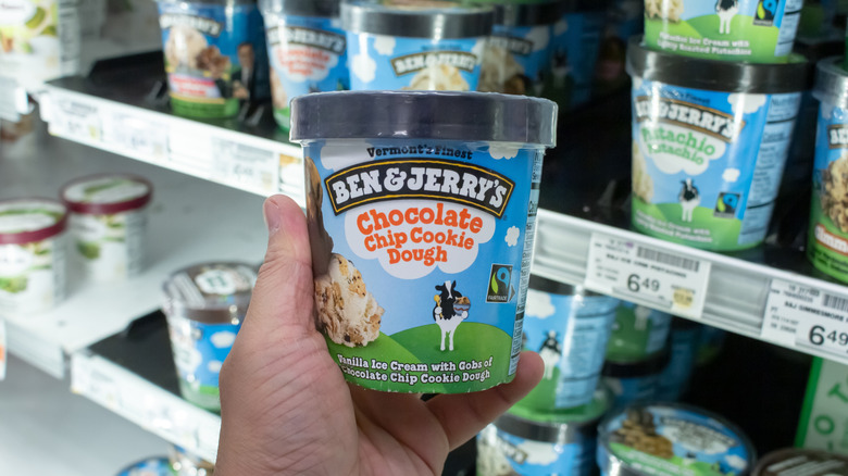 A hand holding a pint of Ben & jerry's chocolate chip cookie dough ice cream in front of an ice cream shelf