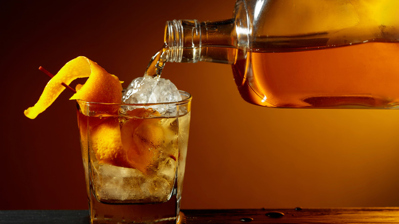 Bourbon being poured from a glear glass bottle into a glass of ice with orange twist