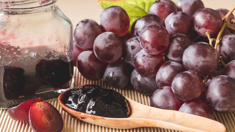 A glass jar of grape jelly with a jellied wooden spoon and a red grape bunch