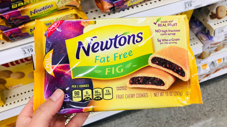 A hand holds up a package of Nabisco Fig Newton Fat Free chewy cookies while shopping at a grocery store.
