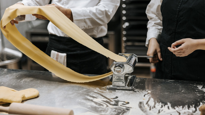 Two chefs work together to use a pasta machine in a professional kitchen
