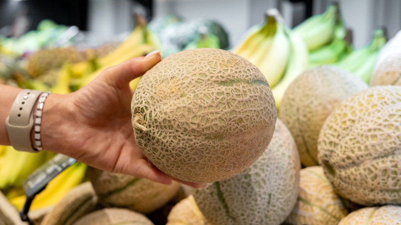 Cantaloupe being held in front of blurred cantaloupe and bananas in the produce section of a store