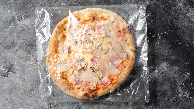 An uncooked frozen pizza still in its plastic wrapper against a slate countertop.