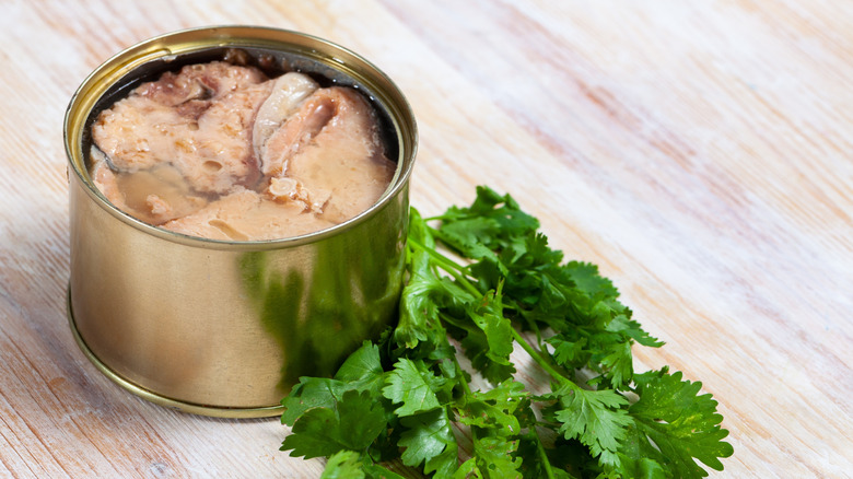An open can of salmon next to some sprigs of parsley on a wooden table.