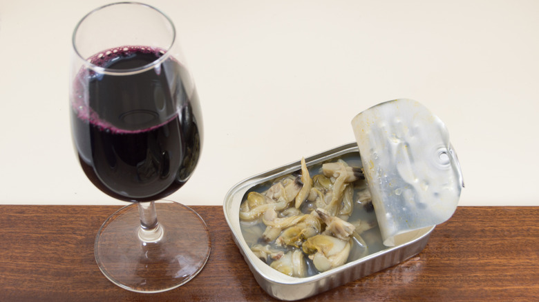 An open tin of clams sits on a table next to a glass of red wine.