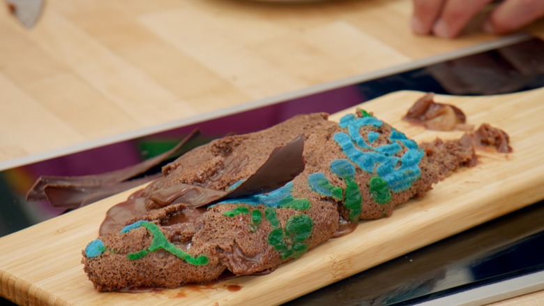 A flat Swiss roll topped with a large chocolate shaving and some green and blue food coloring