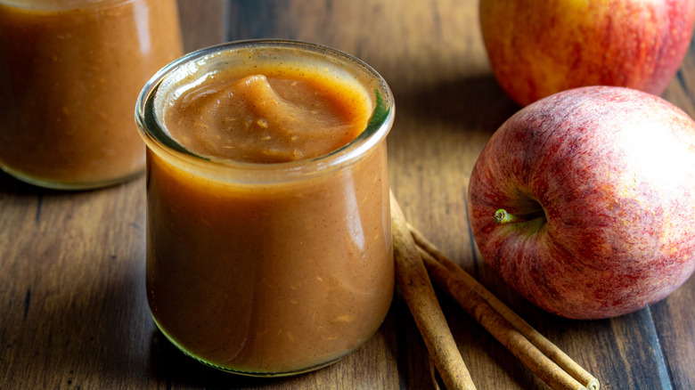 Jars of apple butter beside two whole apples and two cinnamon sticks on a wooden surface
