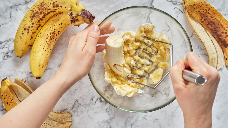 Hands mashing bananas in a bowl with a potato masher
