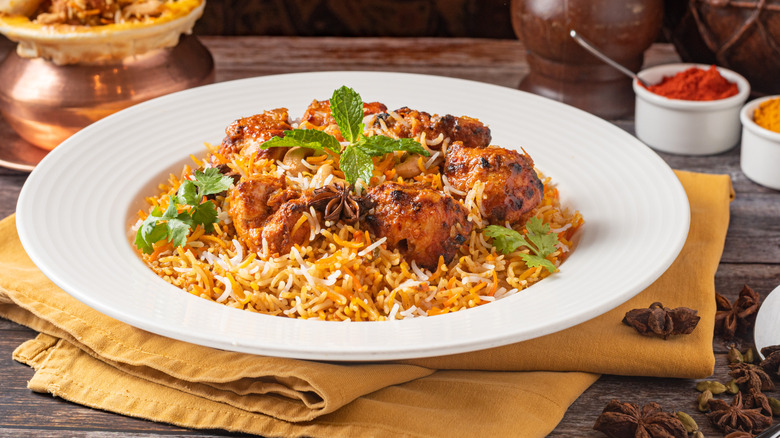 A plate of chicken biryani with a garnish atop a white plate and golden napkin with spices surrounding it.